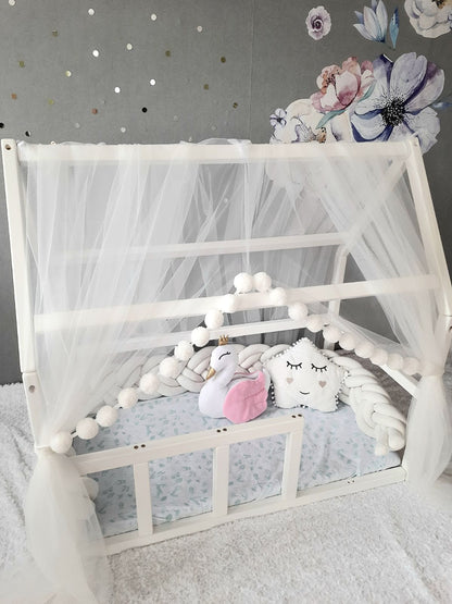 White Montessori Canopy with Pom Poms for nursery. Inside Montessori bed white swan pillow, star pillow and Double braided crib bumper. Front side with a slight view from the top-left angle