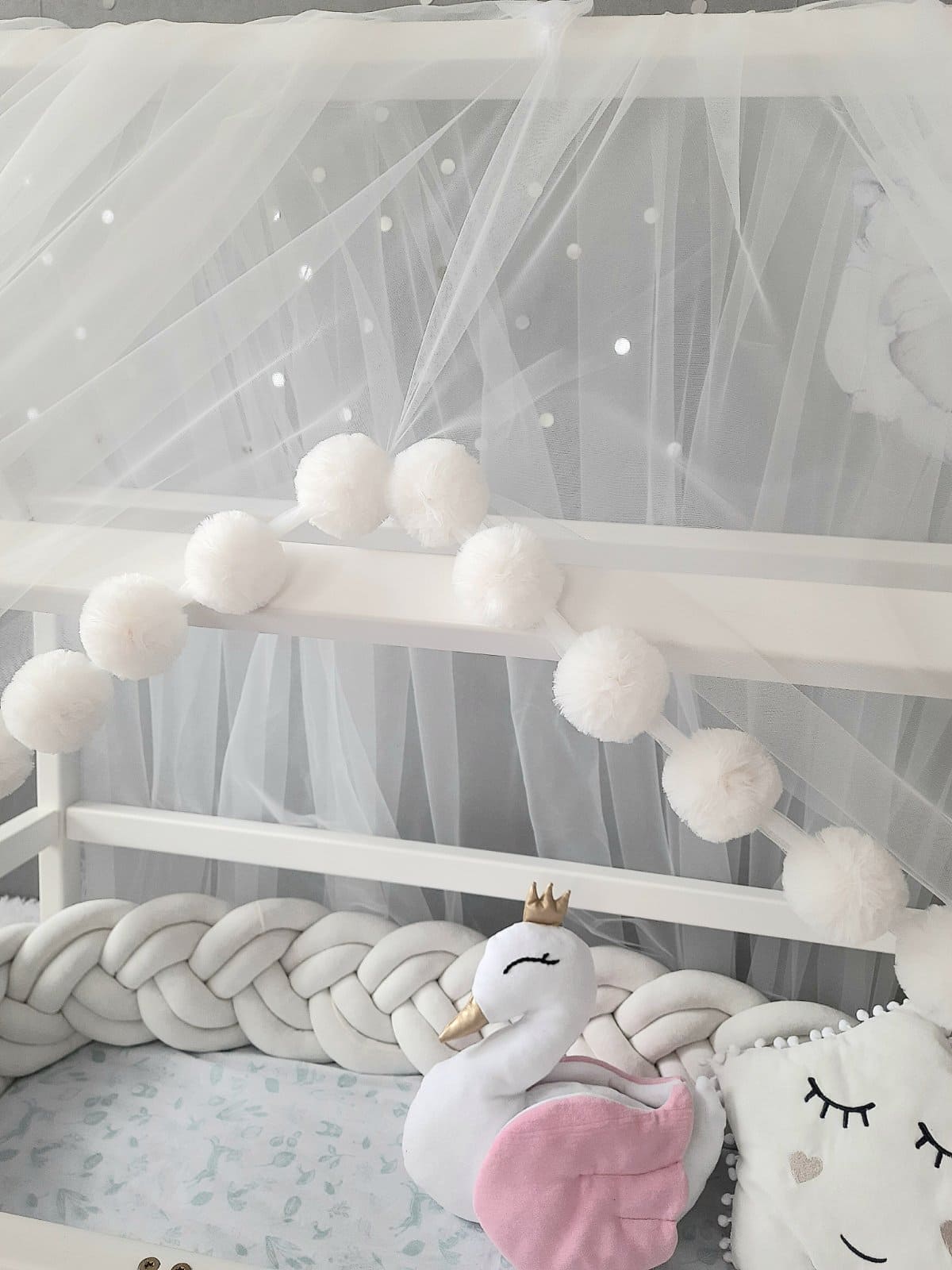 White Montessori Canopy with Pom Poms for nursery. Inside Montessori bed white swan pillow, star pillow and Double braided crib bumper. Close-up slight right view of the front side