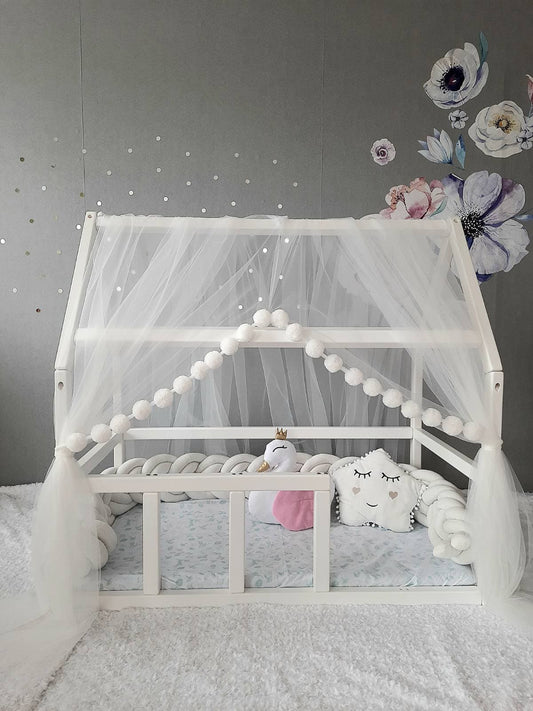 White Montessori Canopy with Pom Poms for nursery. Inside Montessori bed white swan pillow, star pillow and Double braided crib bumper. Front side