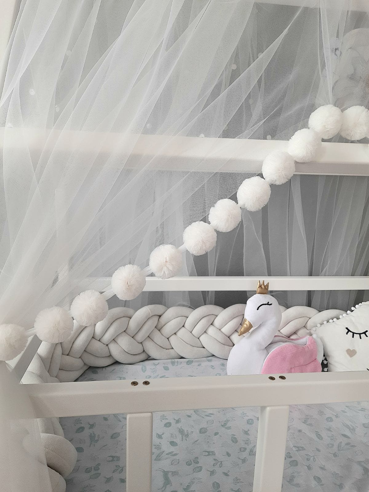 White Montessori Canopy with Pom Poms for nursery. Inside Montessori bed white swan pillow, star pillow and Double braided crib bumper. Close-up slight left view of the front side
