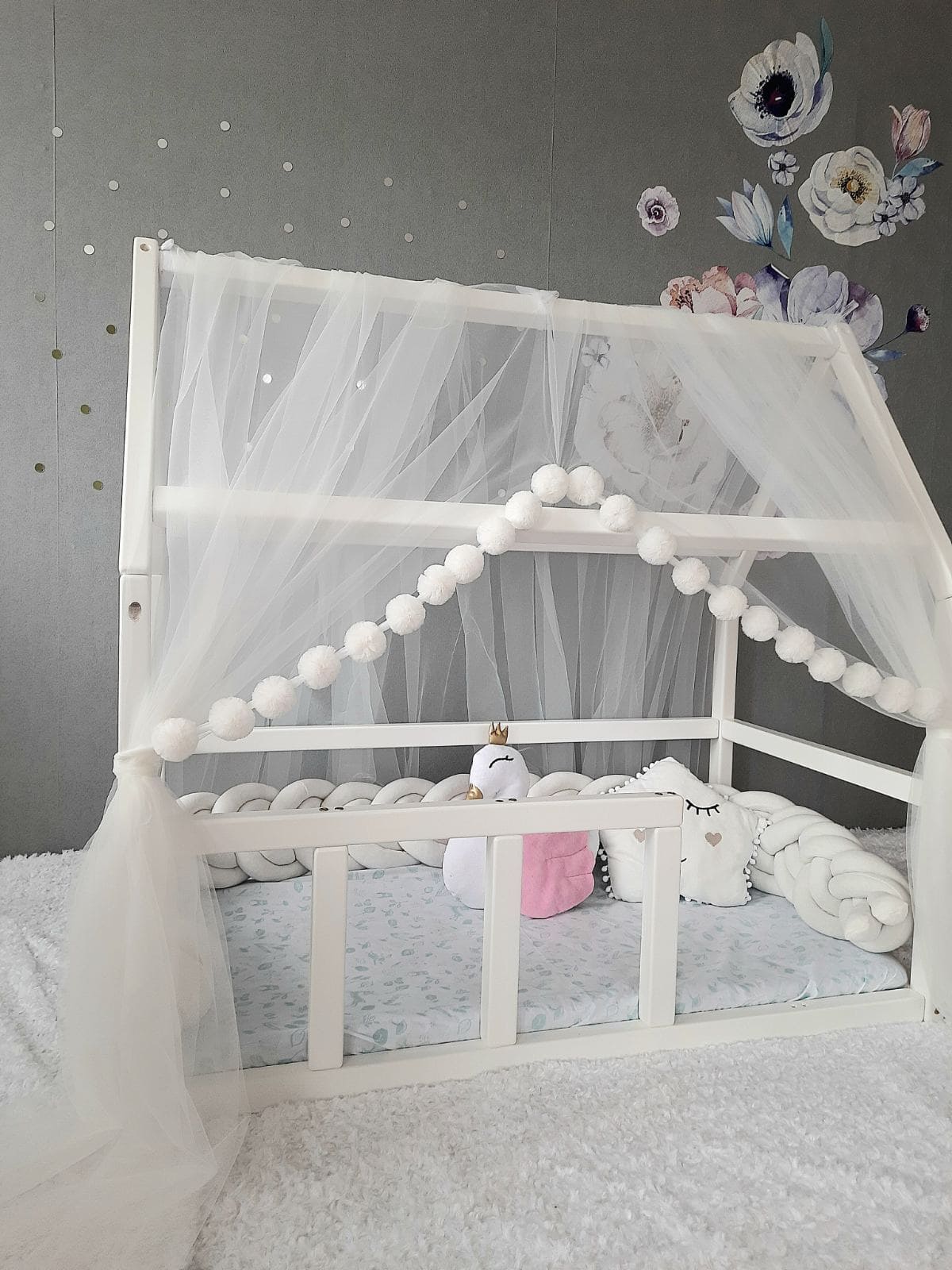 White Montessori Canopy with Pom Poms for nursery. Inside Montessori bed white swan pillow, star pillow and Double braided crib bumper. Front side with a slight view from the left angle