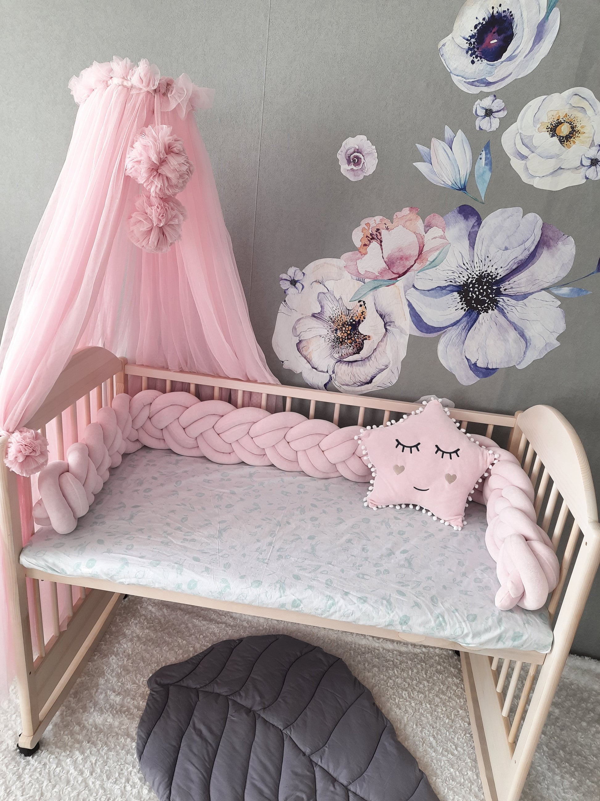 Double braided crib bumper and star pillow on the crib with canopy. Leaf mat on the floor. Light pink color.