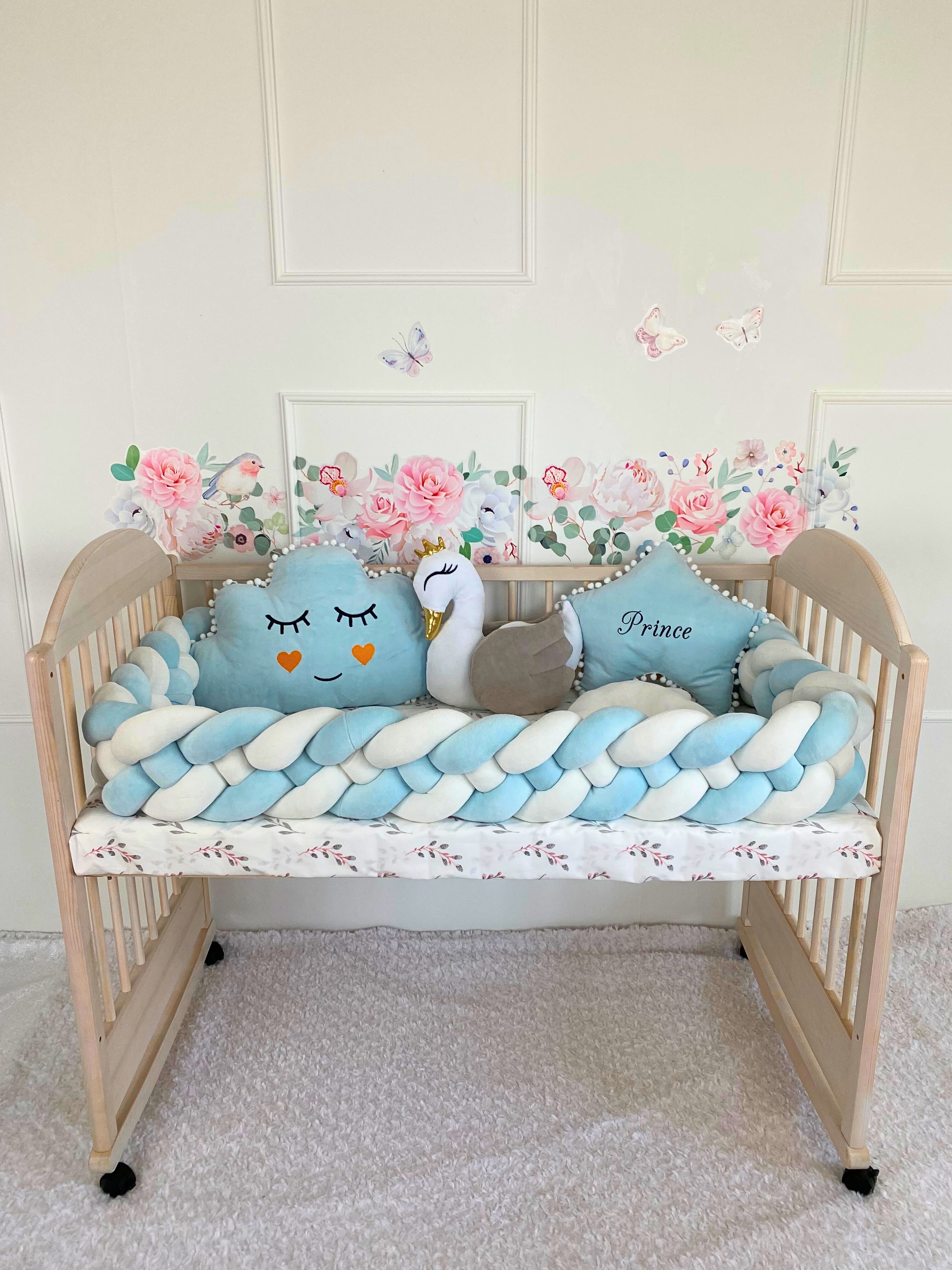 A wooden crib with blue and white braided bumper - weaving in 4 strips, decorative swan and cloud pillows, star pillow with custom  embroidery set against a wall with floral and butterfly stickers.