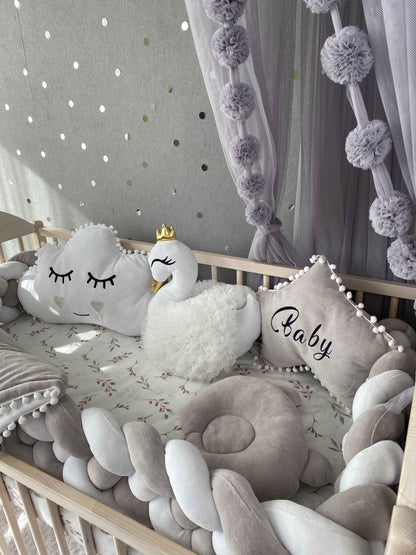 Braided bumper, white cloud and swan pillows, light gray star pillow and orthopedic infant pillow on the crib. 