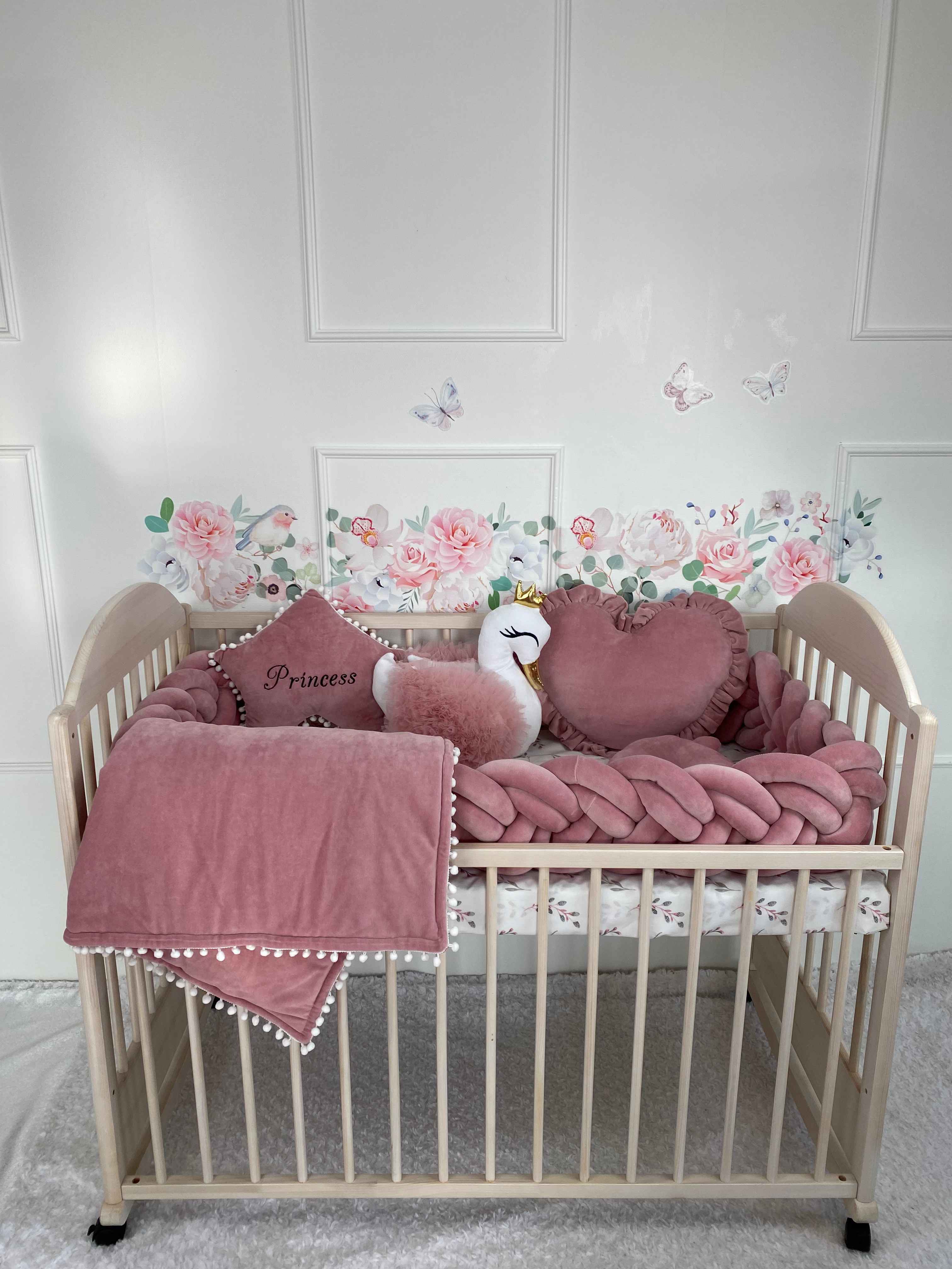 Double braided cot bumper, heart, swan, star pillows and blancet on the wooden crib againt the white wall. Front side