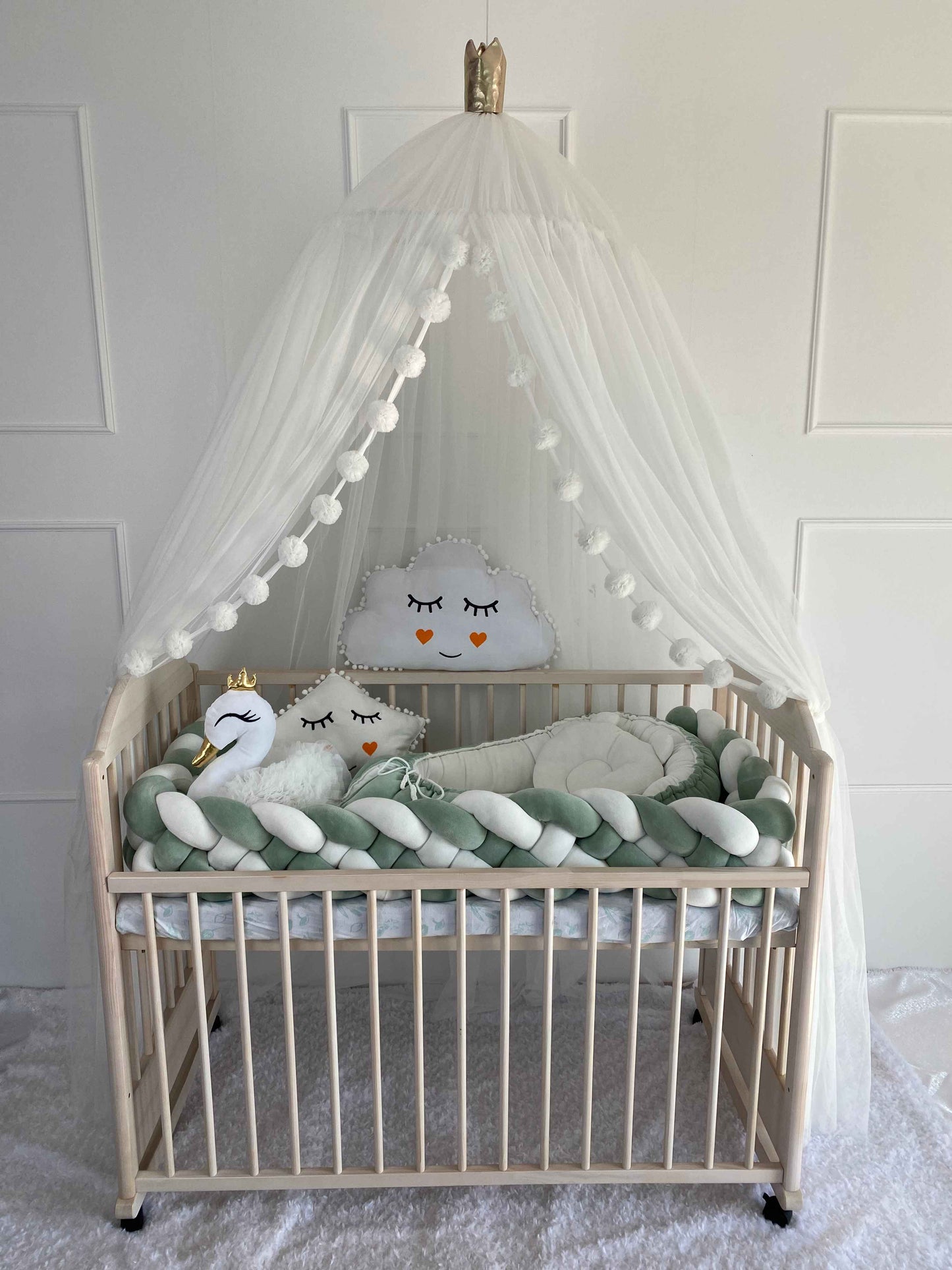 White canopy over wooden crib. Braided cot bumper, baby nest and plush pillows on the crib. Front side