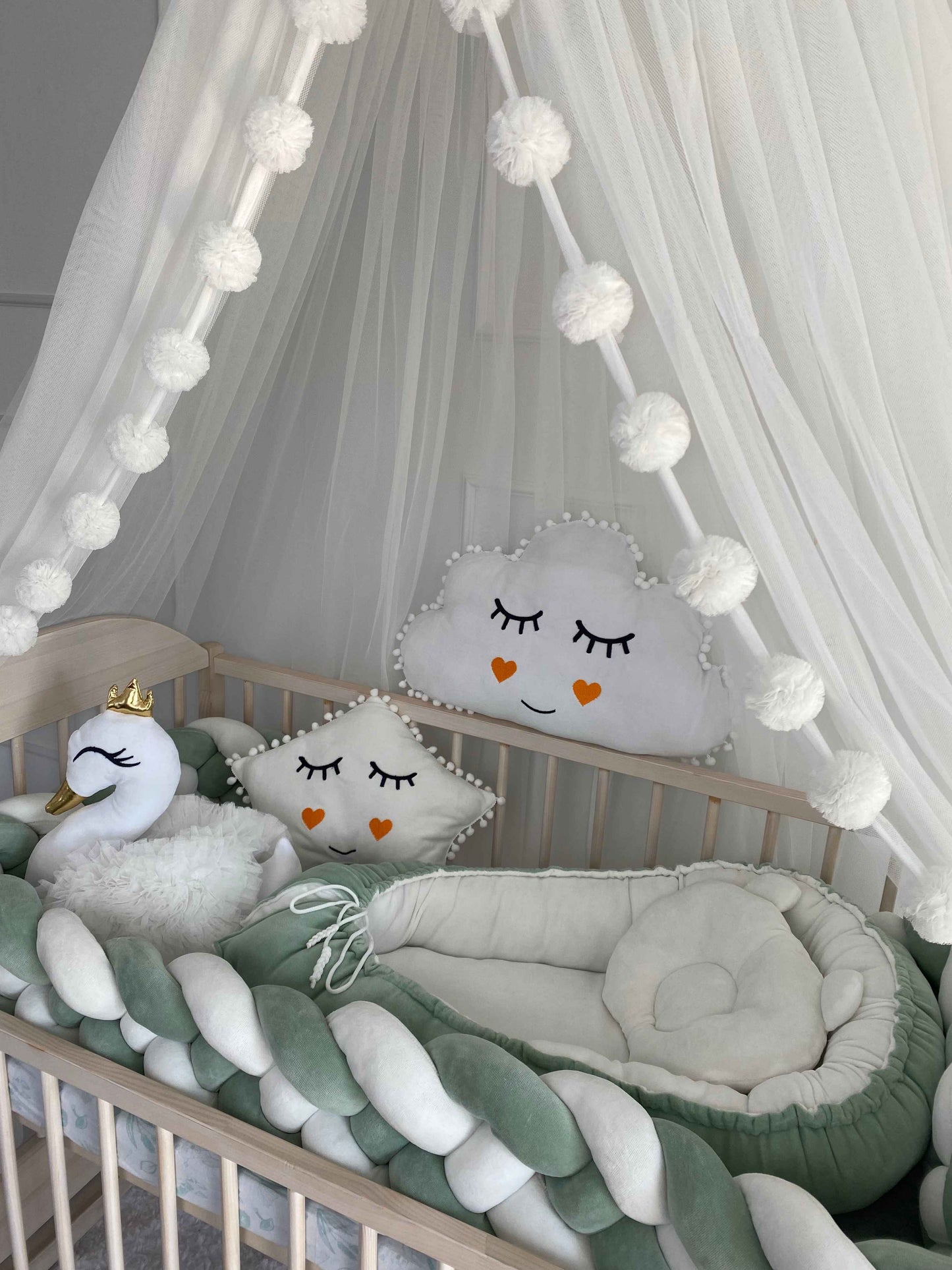 White canopy over wooden crib. Braided cot bumper, baby nest and plush pillows on the crib. Close up view