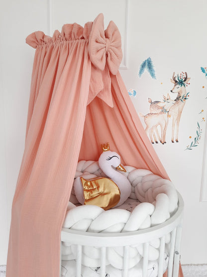 Peach muslin canopy with bow. White braided bumper and swan pillow on the crib