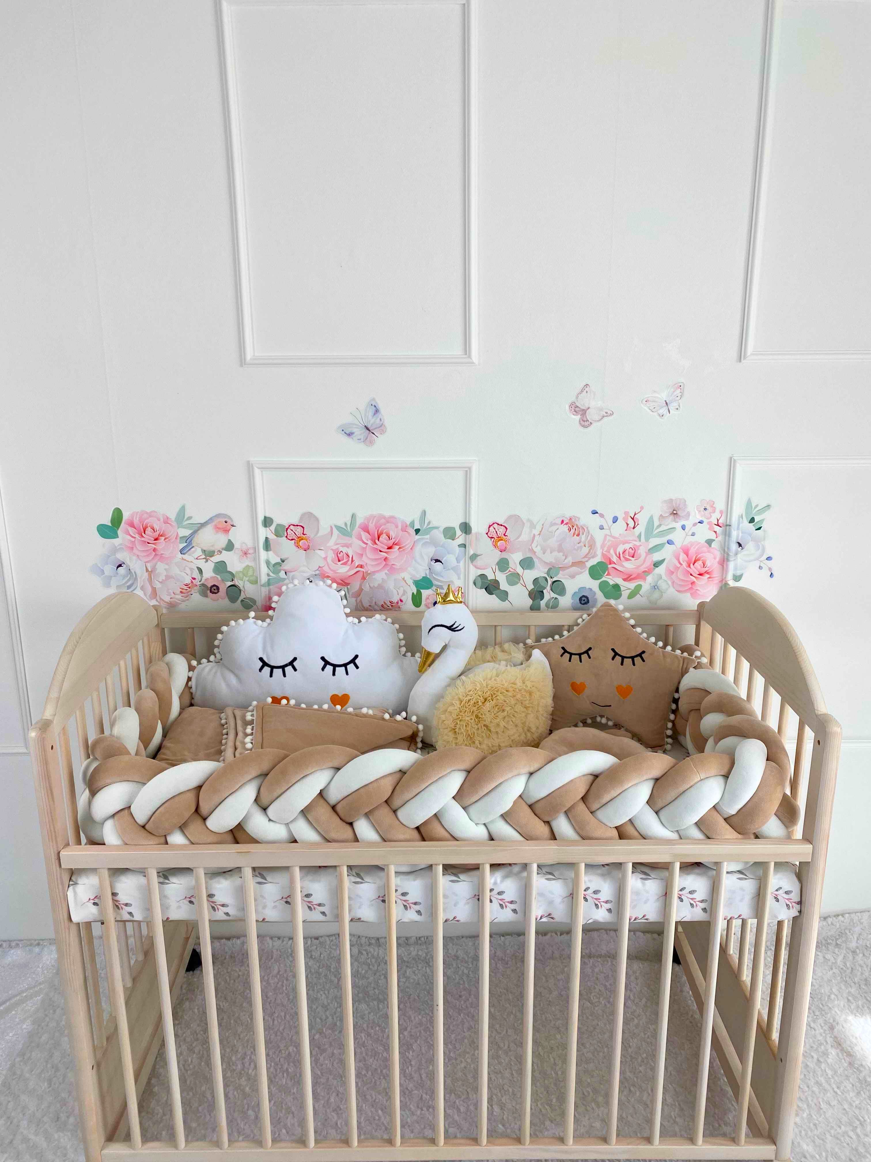 Crib set in cream and white colors. Braided cot bumper and 3 plush pillows on the wooden crib againts the wall. Front side